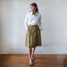 Load image into Gallery viewer, Woman wearing a white shirt and green floral skirt against a white brick wall.