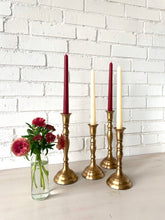 Load image into Gallery viewer, Set of four brass candlesticks with candles against a white brick wall.