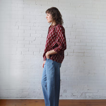 Load image into Gallery viewer, Woman wearing a red patterned blouse and blue jeans against a white brick wall.