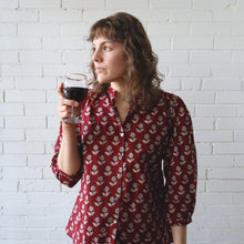 Load image into Gallery viewer, Woman in a red floral blouse holding a glass of red wine against a white brick wall.