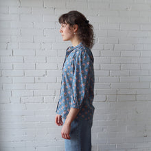 Load image into Gallery viewer, Woman wearing a blue floral blouse against a white brick wall