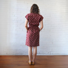 Load image into Gallery viewer, Woman wearing a red patterned dress against a white brick wall.