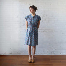 Load image into Gallery viewer, Woman wearing a blue floral dress standing against a white brick wall.