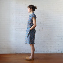 Load image into Gallery viewer, Woman wearing a blue floral dress standing against a white brick wall.