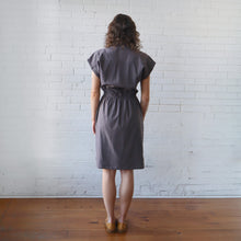 Load image into Gallery viewer, Woman wearing a gray dress standing against a white brick wall.