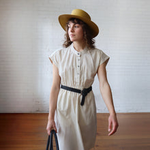 Load image into Gallery viewer, Woman wearing a white dress and yellow hat against a white brick wall.