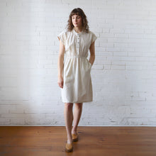 Load image into Gallery viewer, Woman wearing a white dress standing against a white brick wall.