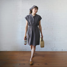 Load image into Gallery viewer, Woman in a gray dress holding a bottle and a woven bag against a white brick wall.