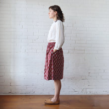 Load image into Gallery viewer, Woman wearing a white blouse and red patterned skirt standing against a white brick wall.