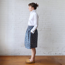 Load image into Gallery viewer, Woman wearing a white blouse and blue patterned skirt against a white brick wall.