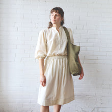 Load image into Gallery viewer, Woman in a beige blouse and skirt holding a green bag against a white brick wall