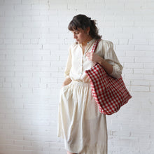 Load image into Gallery viewer, Woman holding a red and white checkered bag against a white brick wall