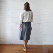 Load image into Gallery viewer, Person wearing a white blouse and gray skirt standing against a white brick wall.