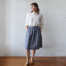 Load image into Gallery viewer, Woman wearing a white shirt and blue skirt standing against a white brick wall.