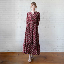 Load image into Gallery viewer, Woman wearing a long burgundy dress with a floral pattern against a white brick wall.