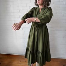 Load image into Gallery viewer, Woman wearing an olive green dress against a white brick wall.