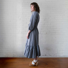 Load image into Gallery viewer, Woman wearing a blue checkered dress standing against a white brick wall.