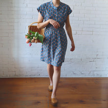 Load image into Gallery viewer, Woman in a blue floral dress holding a basket of flowers against a white brick wall.
