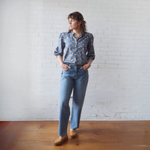 Load image into Gallery viewer, Person wearing a patterned shirt and blue jeans standing against a white brick wall.

