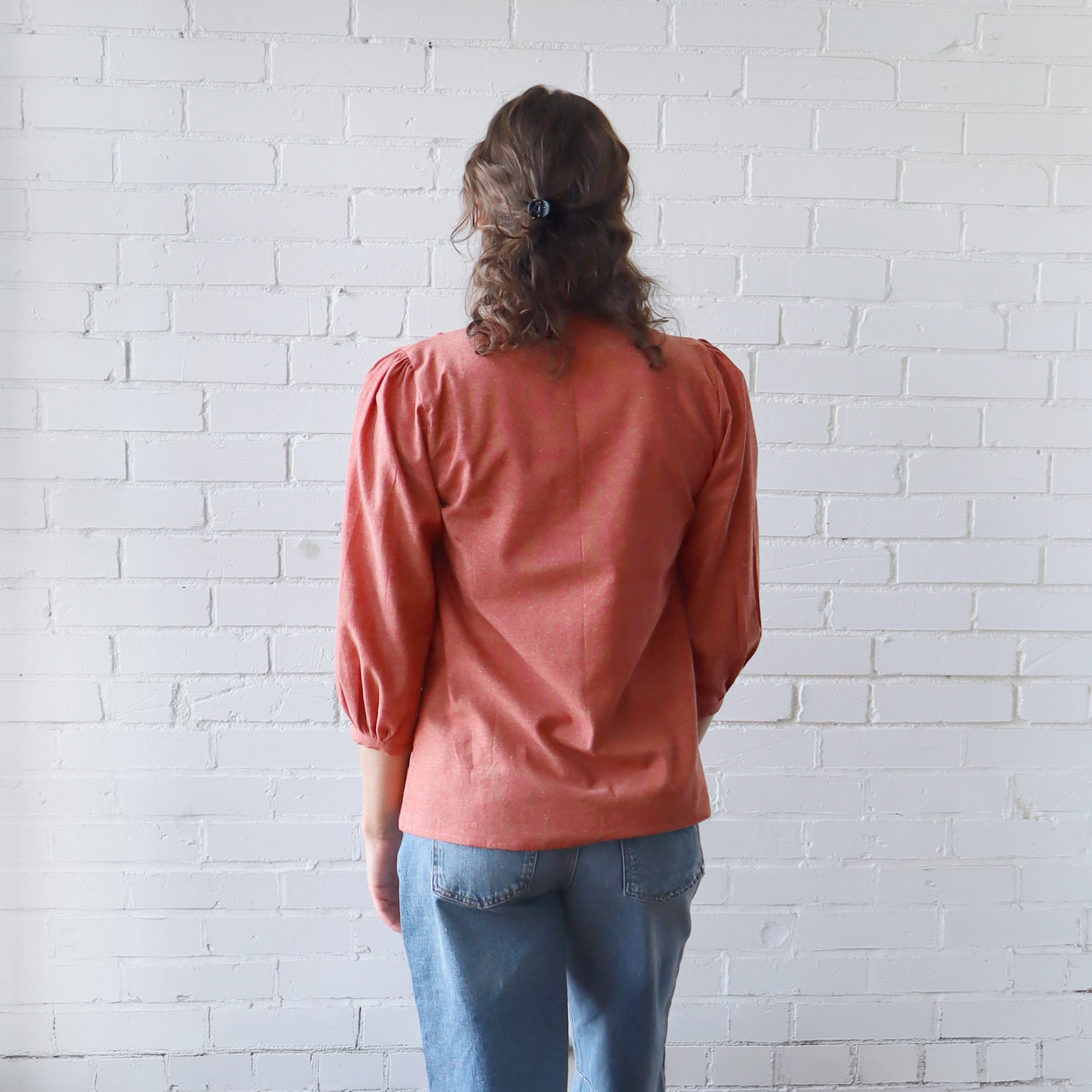 Person wearing a pink blouse and blue jeans standing against a white brick wall.