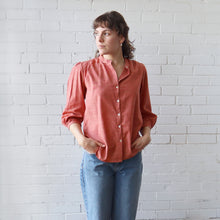 Load image into Gallery viewer, Woman wearing a coral shirt and blue jeans against a white brick wall
