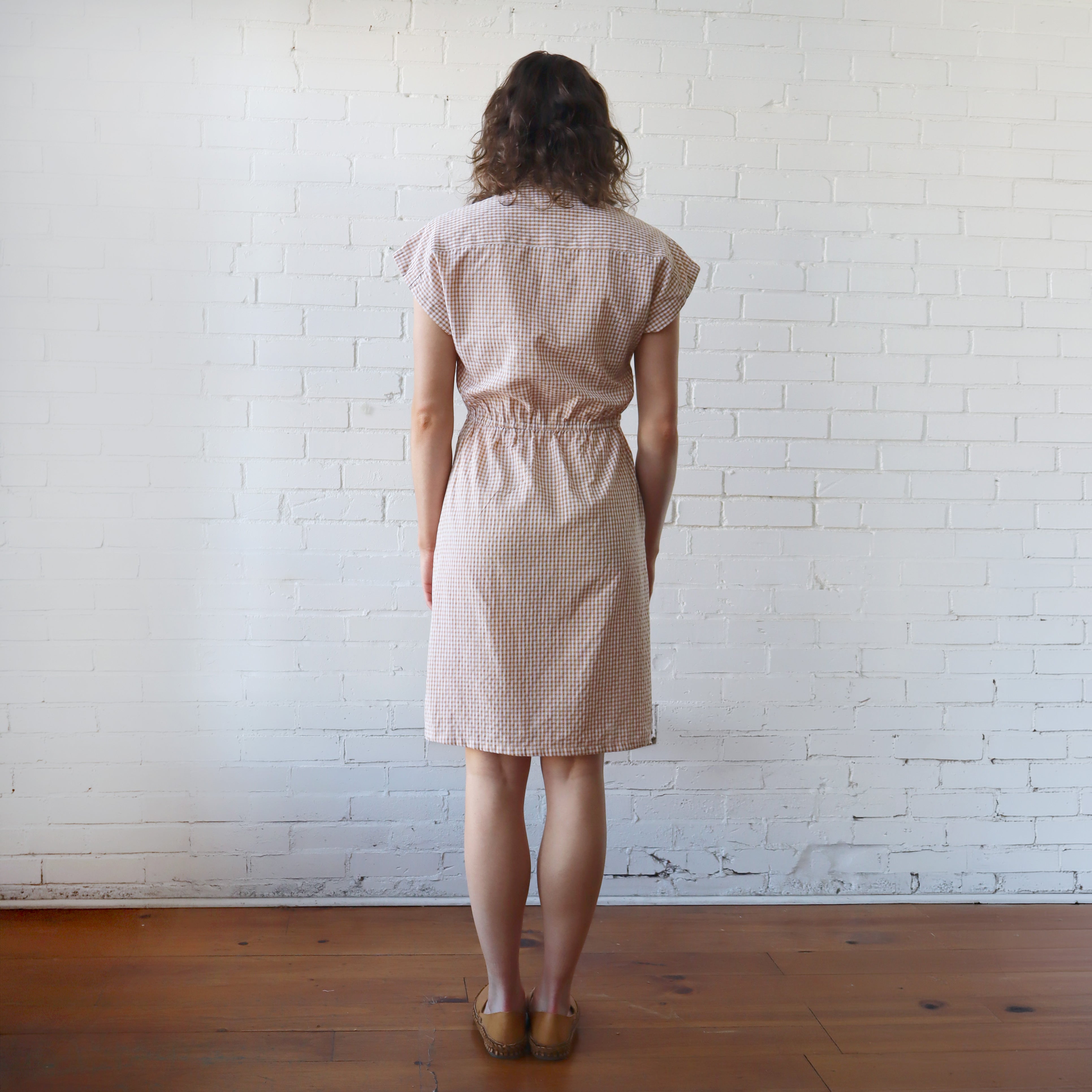Woman wearing a beige checkered dress standing against a white brick wall.