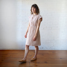 Load image into Gallery viewer, Woman wearing a pink checkered dress standing against a white brick wall.
