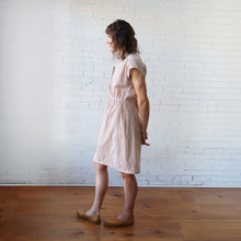 Load image into Gallery viewer, Woman wearing a pink checkered dress standing against a white brick wall.
