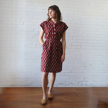 Load image into Gallery viewer, Woman wearing a red patterned dress standing against a white brick wall.
