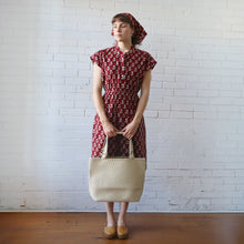 Load image into Gallery viewer, Woman in a red patterned dress holding a woven bag against a white brick wall.
