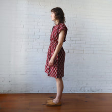 Load image into Gallery viewer, Woman wearing a red patterned dress standing against a white brick wall.

