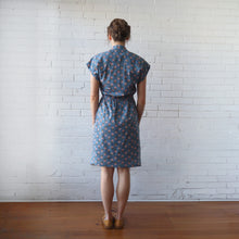 Load image into Gallery viewer, Woman wearing a blue patterned dress standing against a white brick wall.
