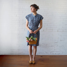 Load image into Gallery viewer, Woman in a blue patterned dress holding a basket of flowers against a white brick wall.
