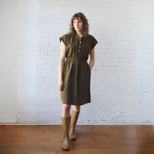 Load image into Gallery viewer, Woman wearing a green dress and brown boots standing against a white brick wall.
