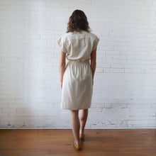 Load image into Gallery viewer, Woman wearing a beige dress standing against a white brick wall.
