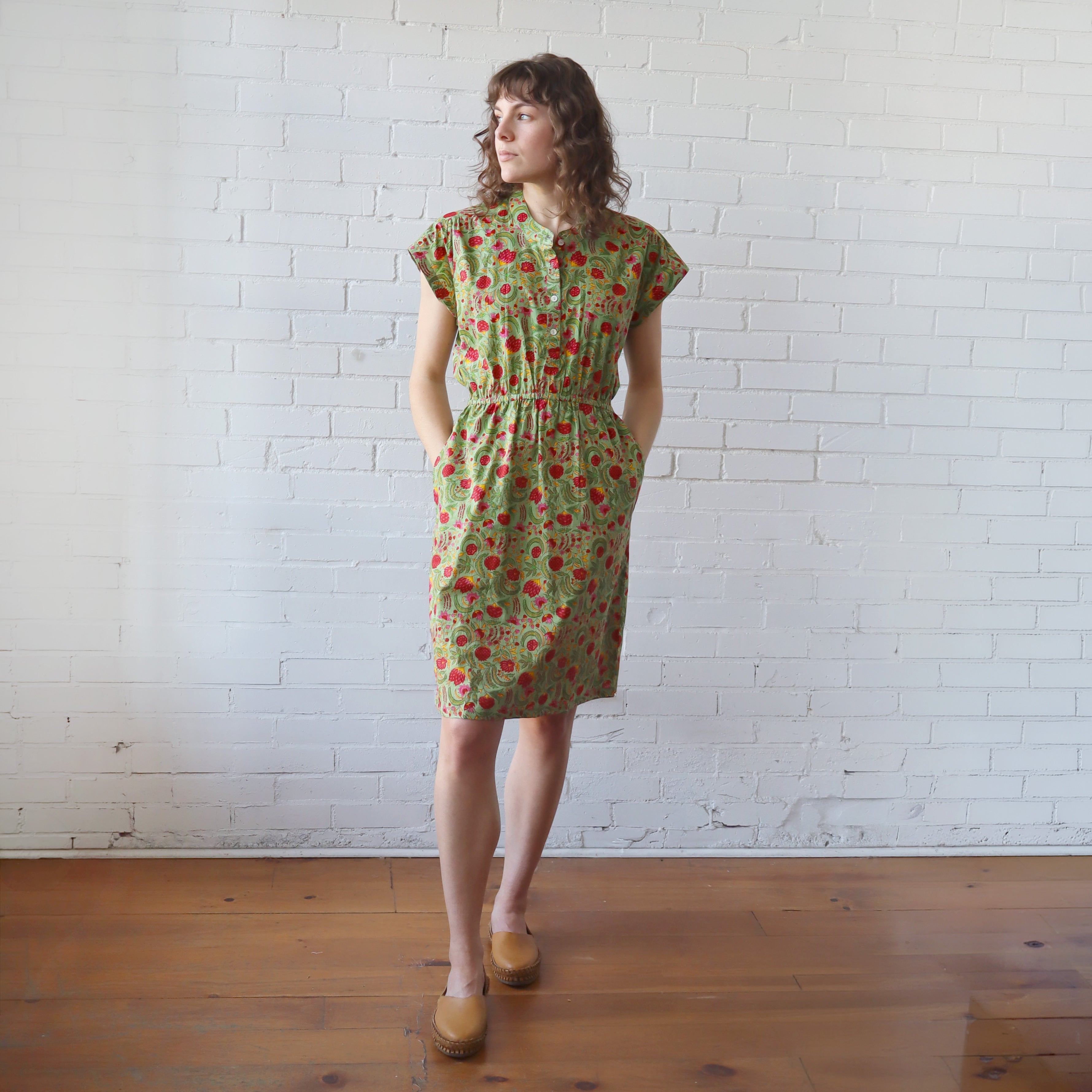 Woman wearing a green floral dress standing against a white brick wall.