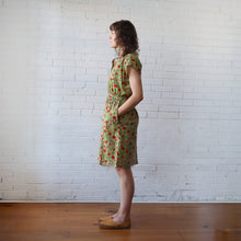Load image into Gallery viewer, Woman wearing a green floral dress standing against a white brick wall.
