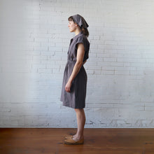 Load image into Gallery viewer, Woman wearing a gray dress and headscarf standing against a white brick wall.
