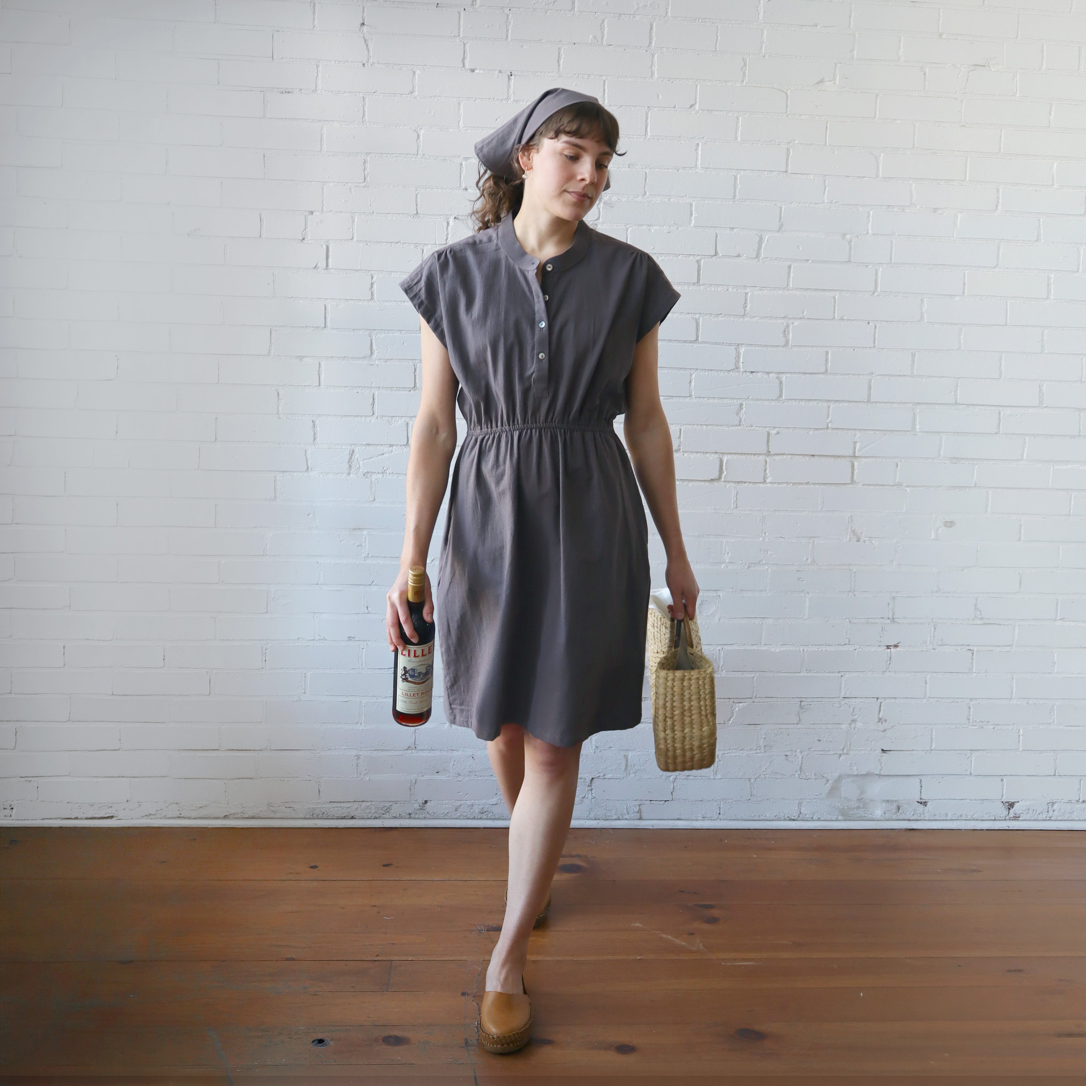 Woman in a gray dress holding a bottle and a woven bag against a white brick wall.