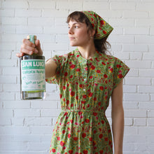 Load image into Gallery viewer, Woman in a green floral dress holding a bottle against a white brick wall
