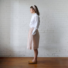 Load image into Gallery viewer, Woman in a white shirt and pink skirt standing against a white brick wall.
