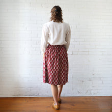 Load image into Gallery viewer, Person wearing a white blouse and red patterned skirt standing against a white brick wall.
