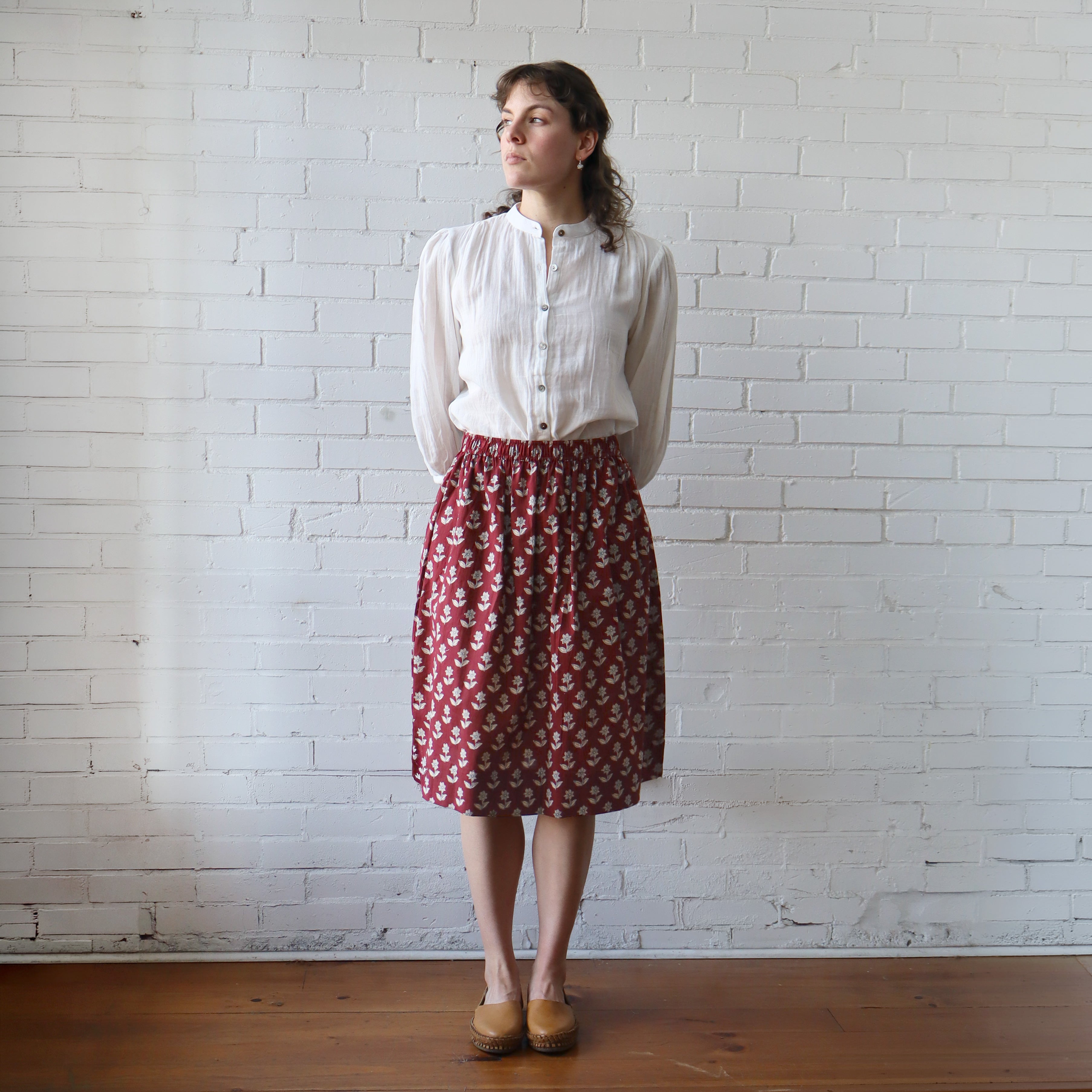 Woman wearing a white blouse and red patterned skirt against a white brick wall.