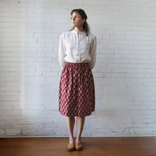 Load image into Gallery viewer, Woman wearing a white blouse and red patterned skirt against a white brick wall.
