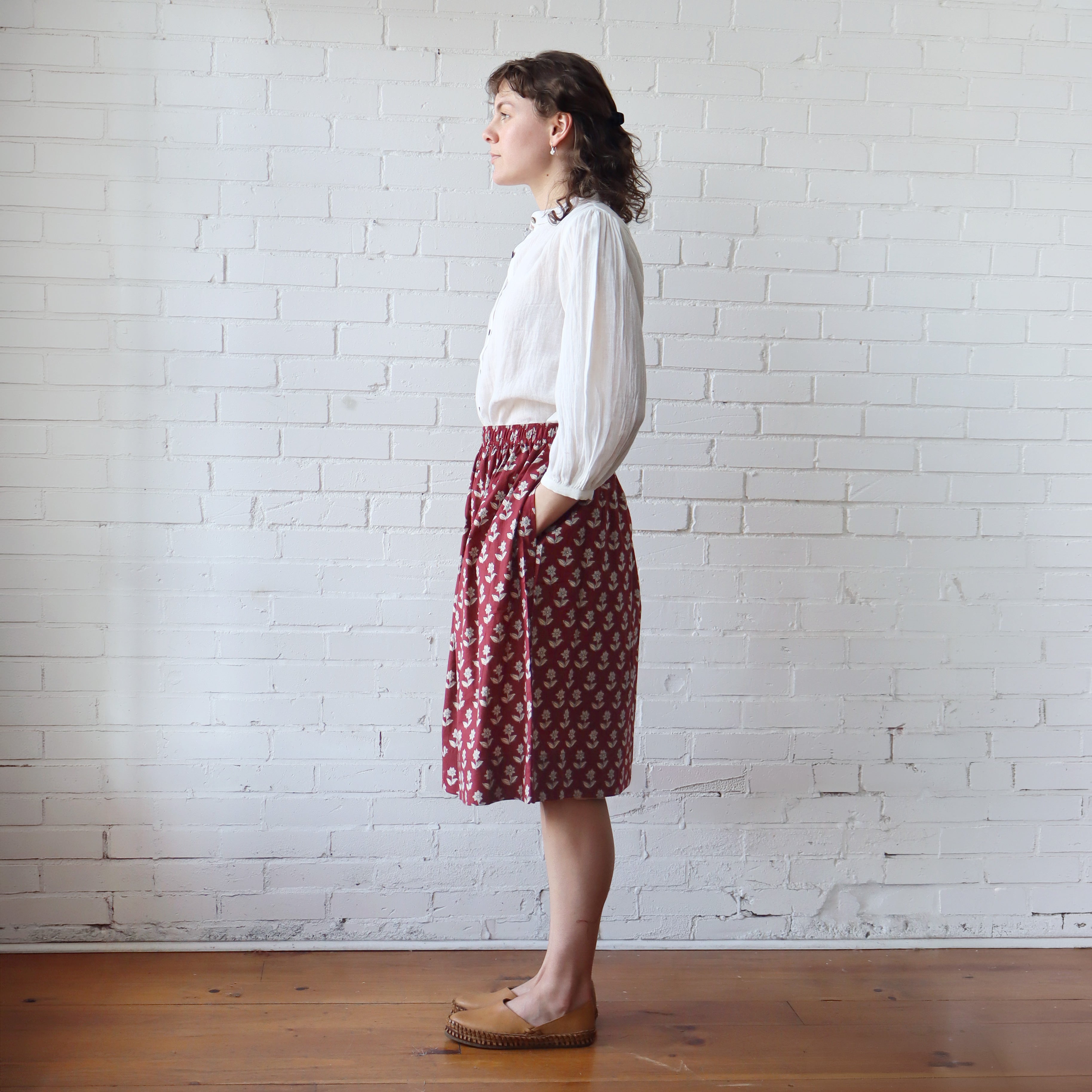 Woman wearing a white blouse and red patterned skirt standing against a white brick wall.
