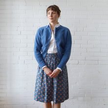 Load image into Gallery viewer, Woman wearing a blue cardigan and patterned skirt against a white brick wall.
