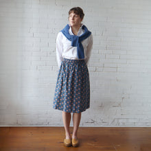 Load image into Gallery viewer, Woman wearing a white blouse, blue patterned skirt, and blue scarf against a white brick wall.
