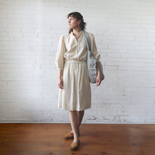 Load image into Gallery viewer, Woman in a beige blouse and skirt holding a striped bag against a white brick wall.
