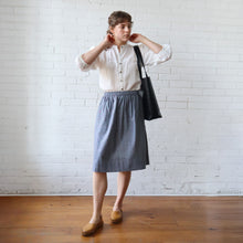 Load image into Gallery viewer, Person wearing a white shirt and gray skirt with a black bag against a white brick wall.

