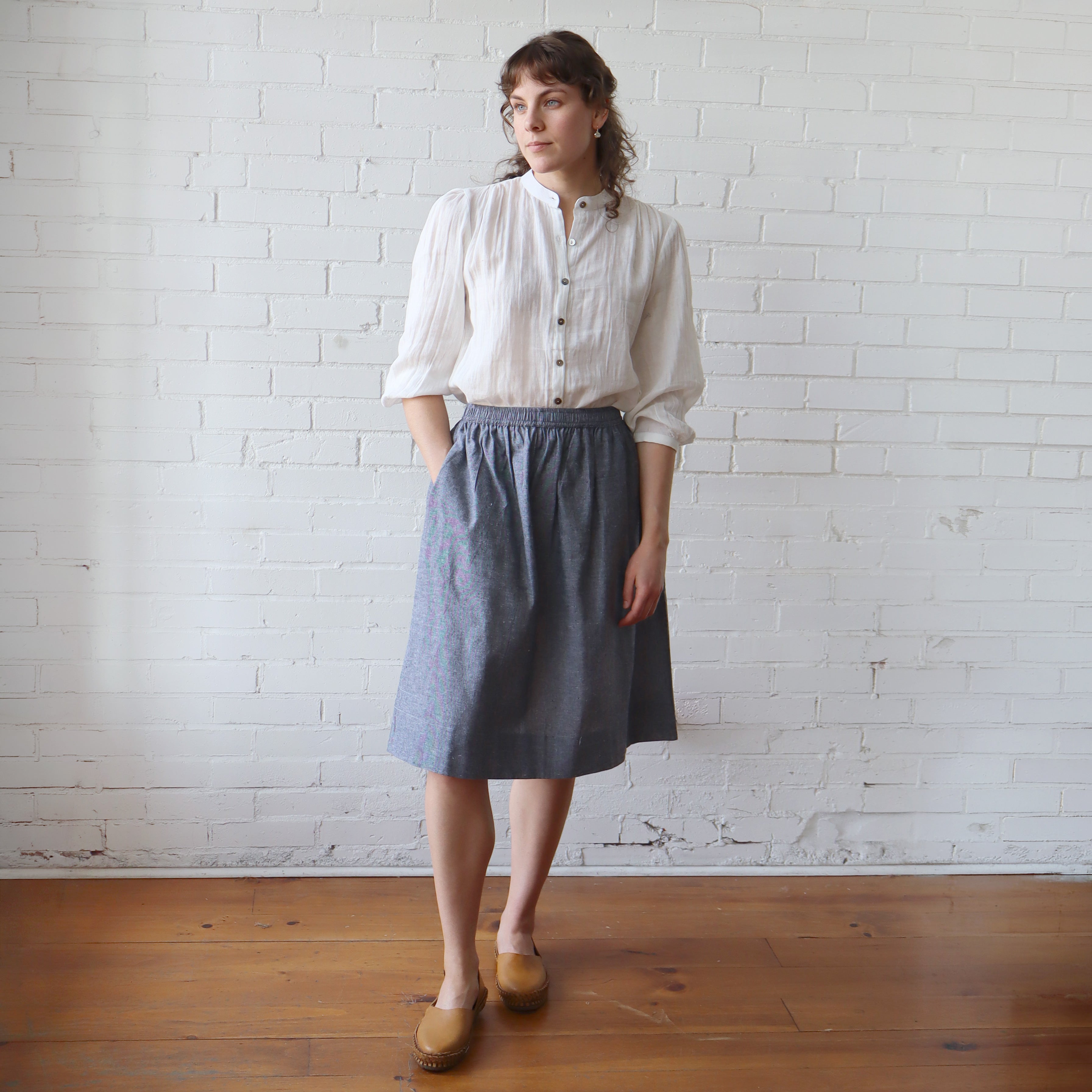 Woman wearing a white blouse and gray skirt standing against a white brick wall.